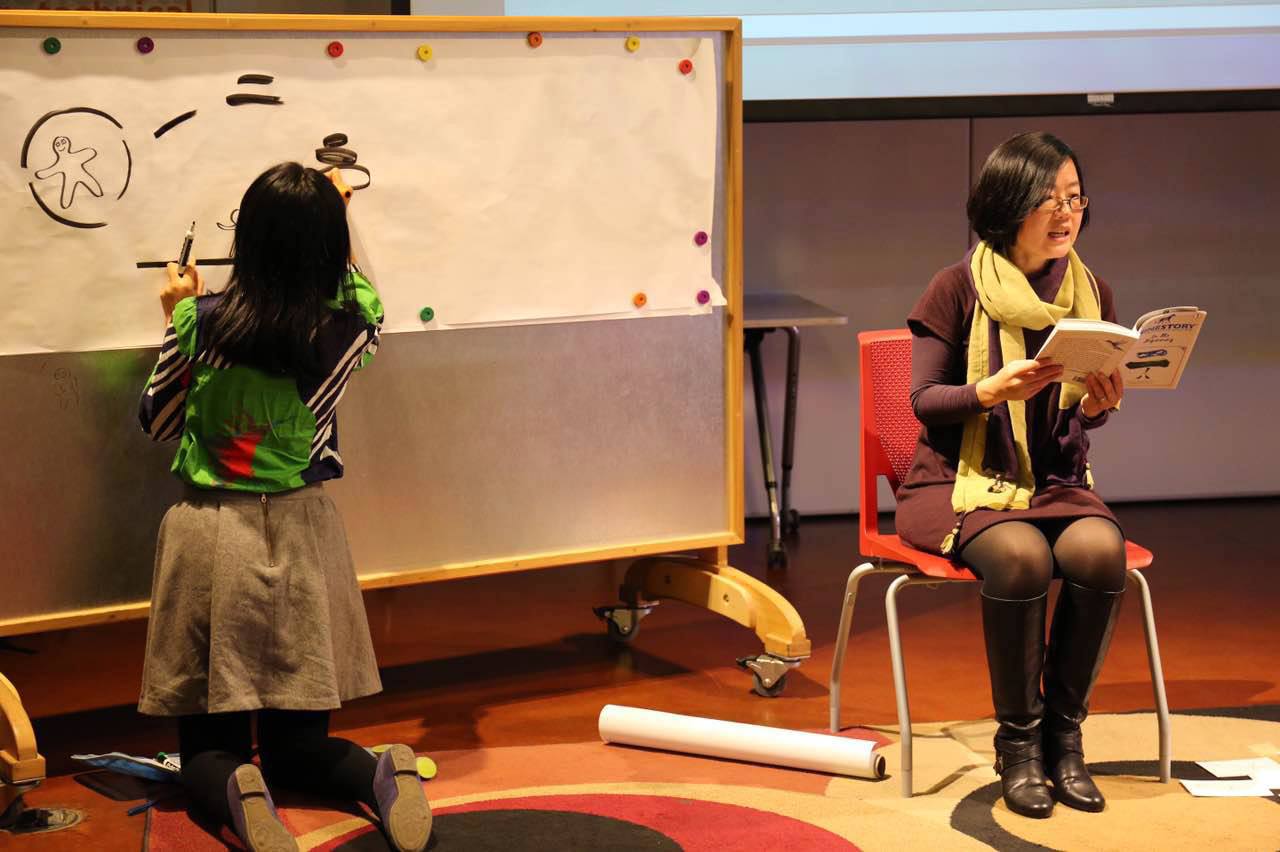 Asian woman sitting in a chair reading out loud. Child facing a white board writing on it.