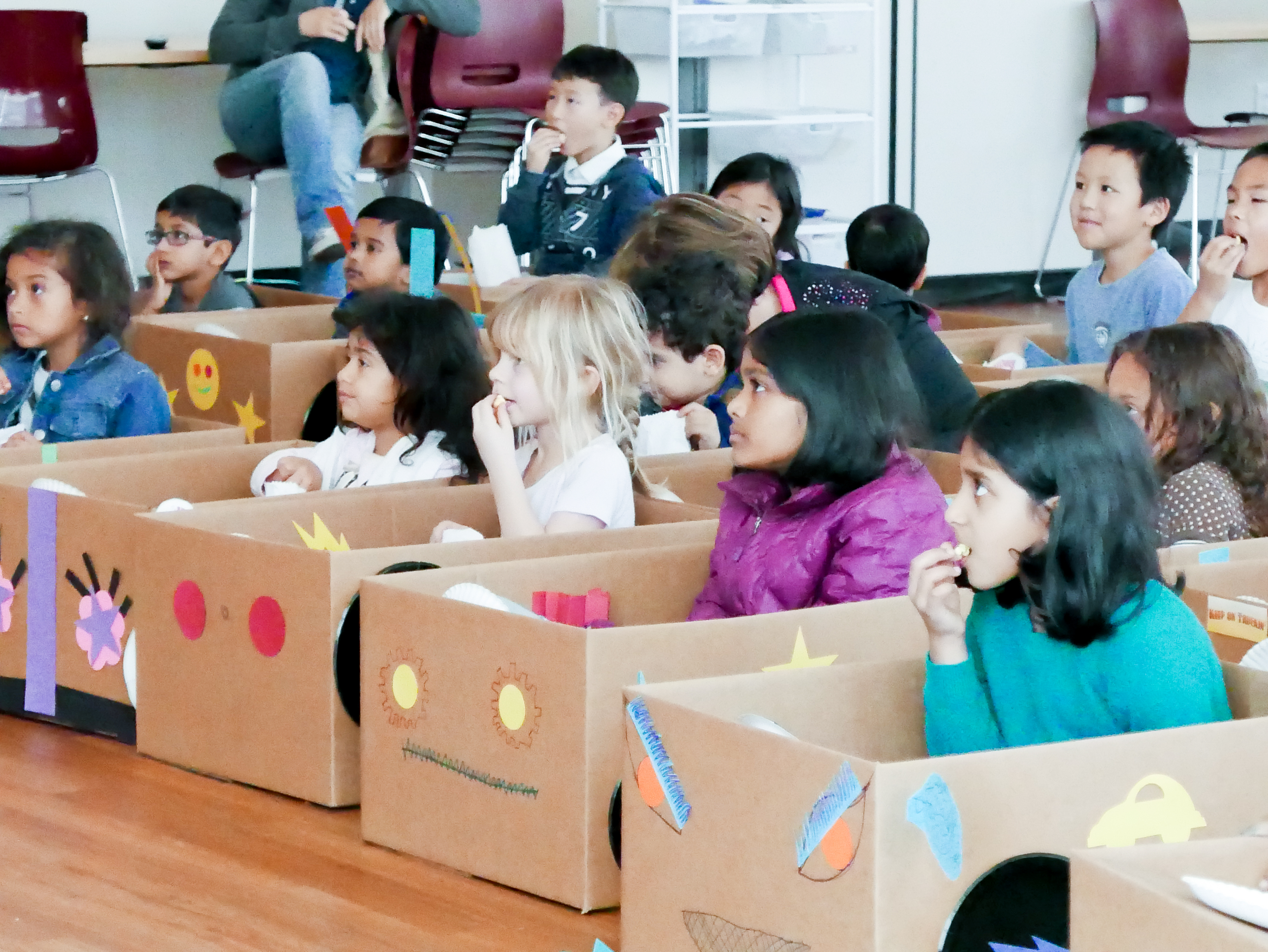 Several kids sitting in home-made cars. The cars are made from cardboard boxes that are decorated. 
