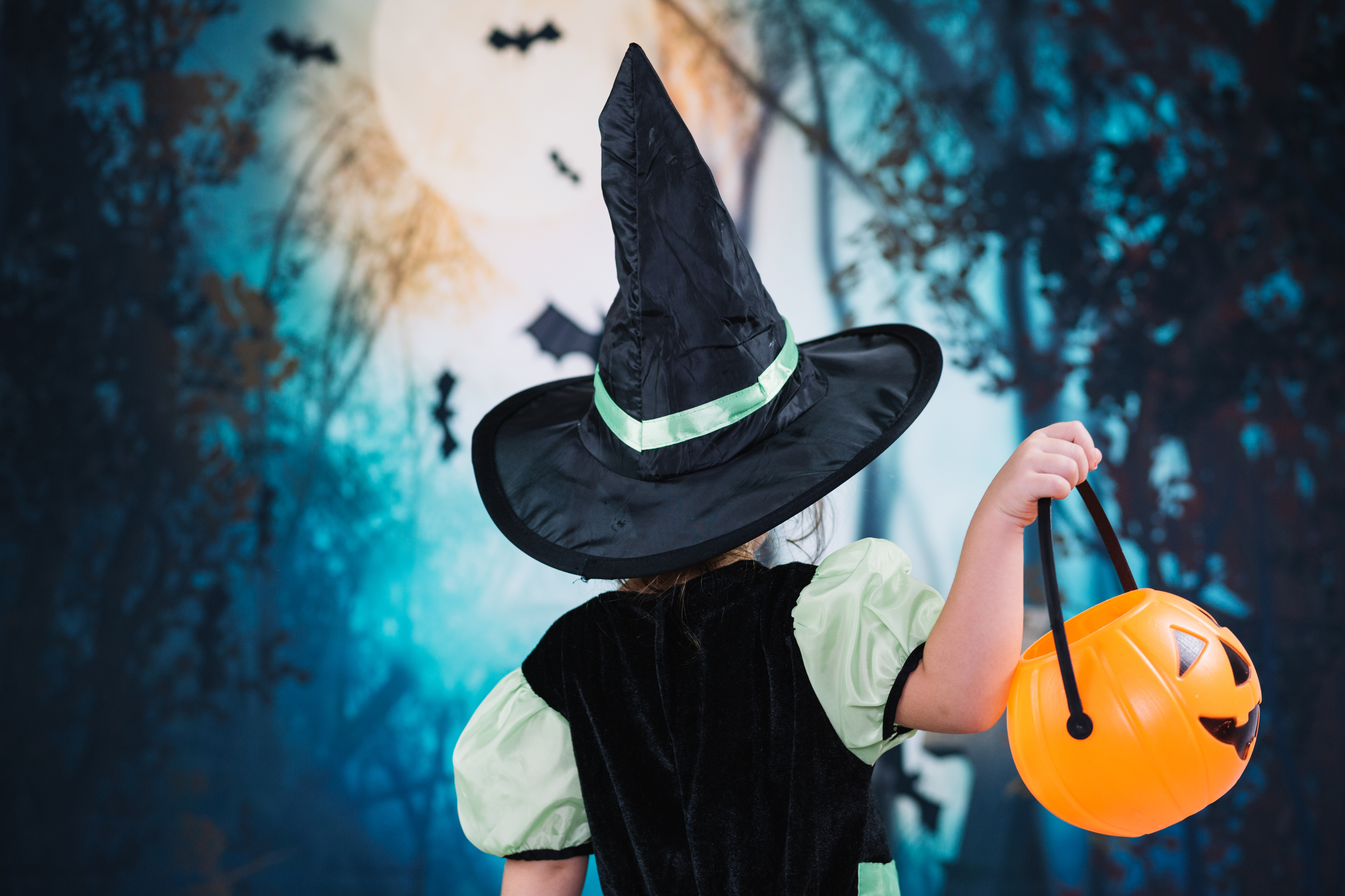 A girl dressed as a witch with her back to the camera. She's holding a jack-o-lantern basket.