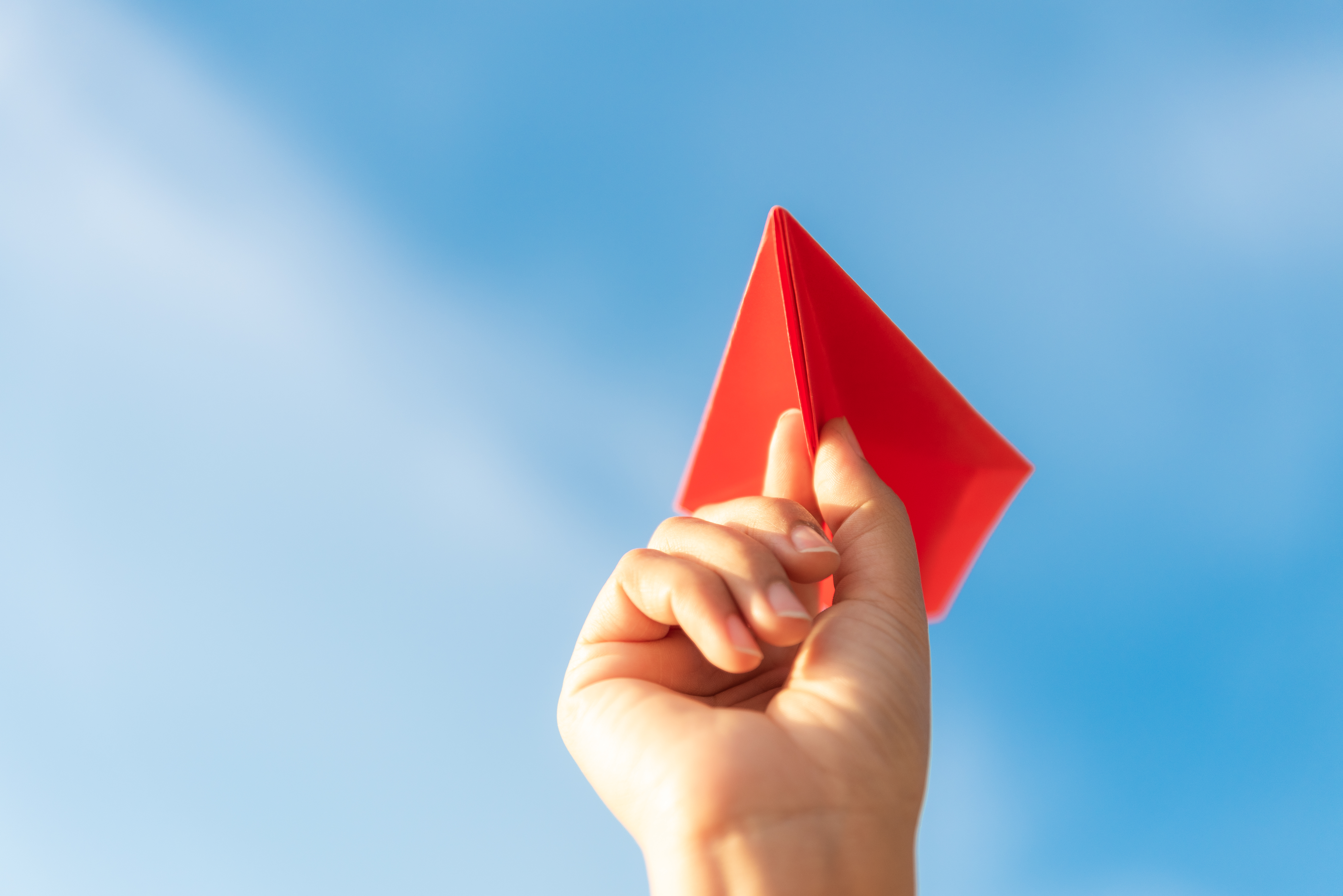 Hand holding a red paper airplane against a blue sky with wispy clouds.