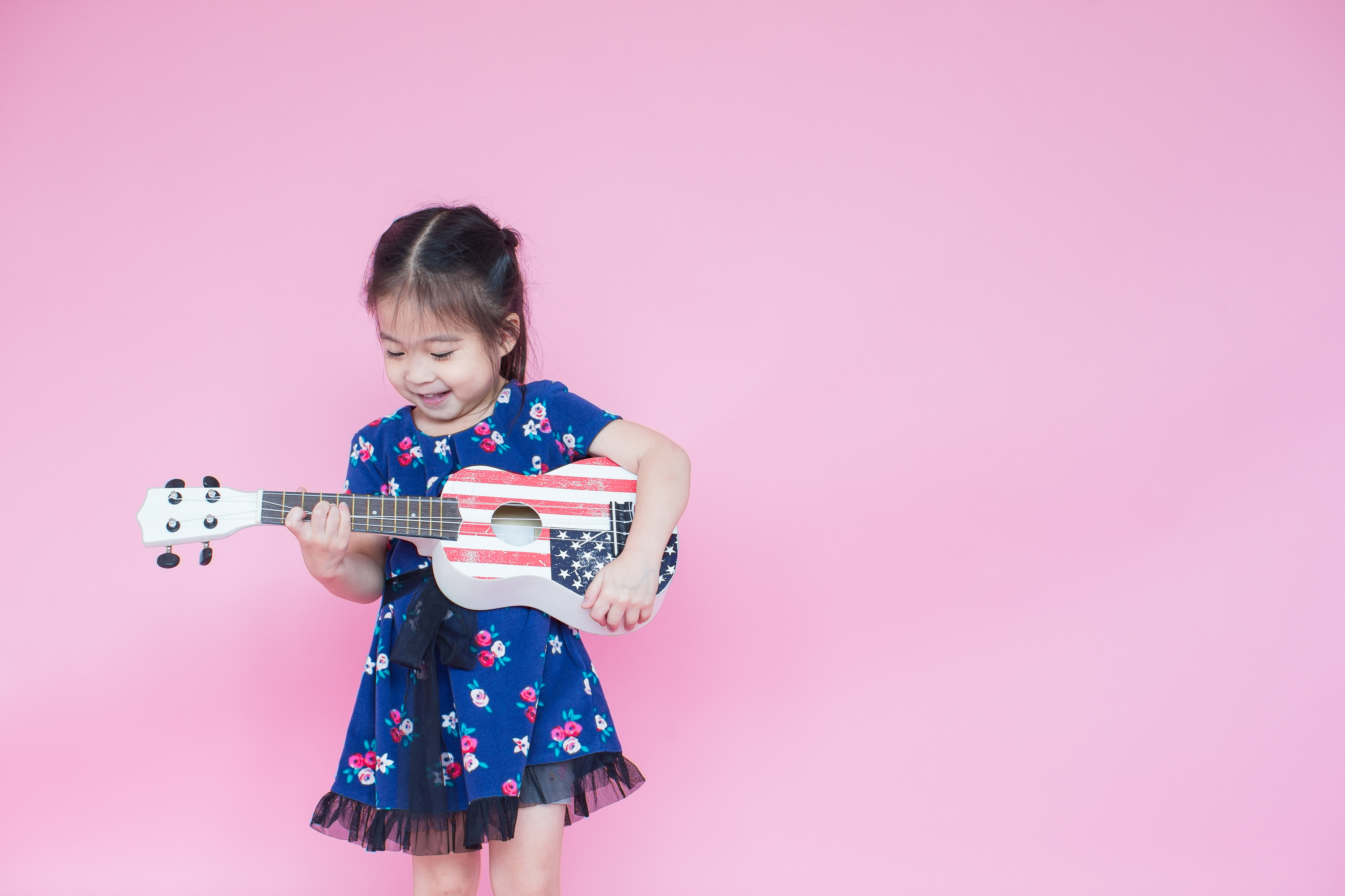 Asian girl wearing a blue dress holding a small guitar with an American flag on it in front of a pink background.