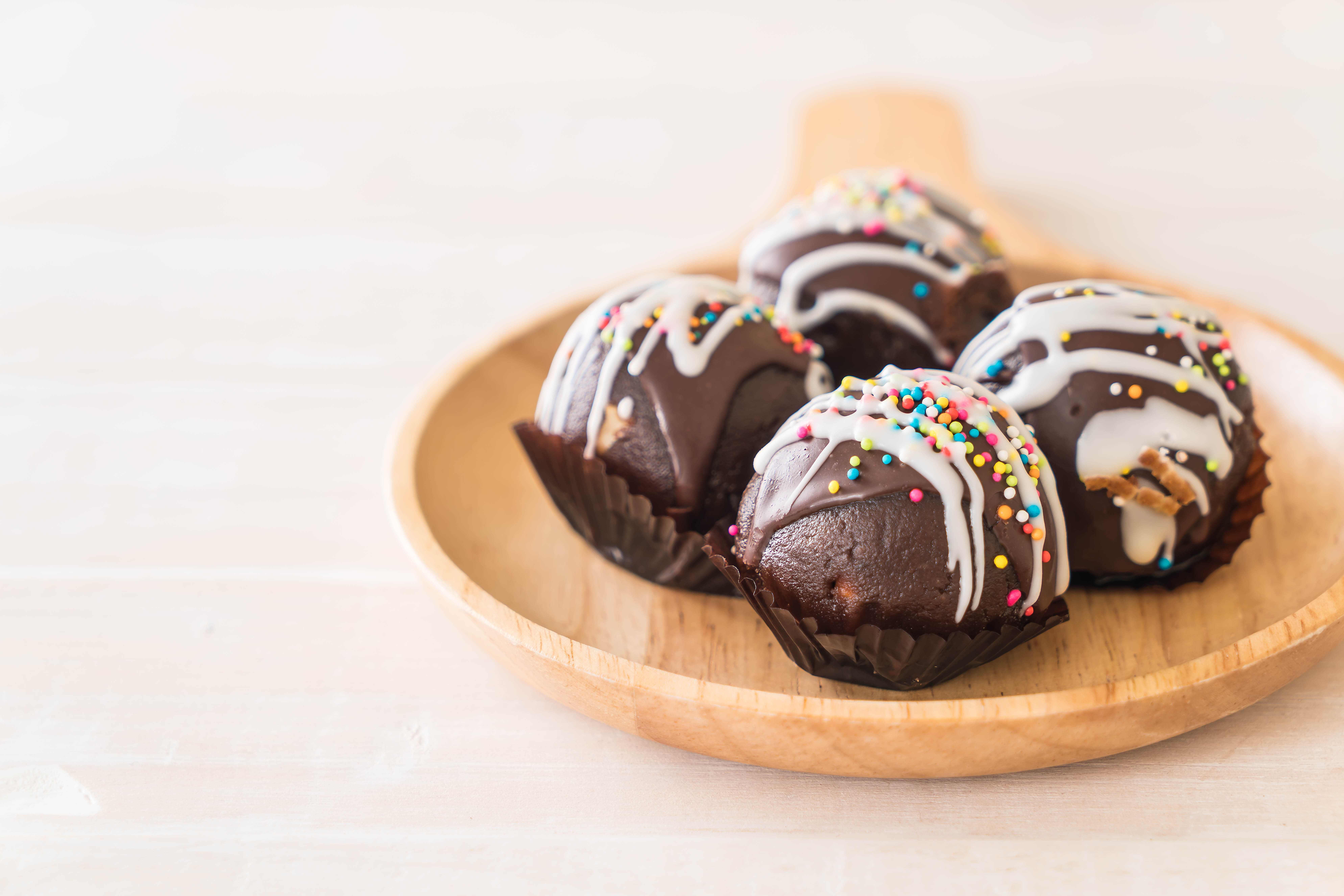Four chocolate truffles with drizzled white icing on a wooden plate in front of a white background.