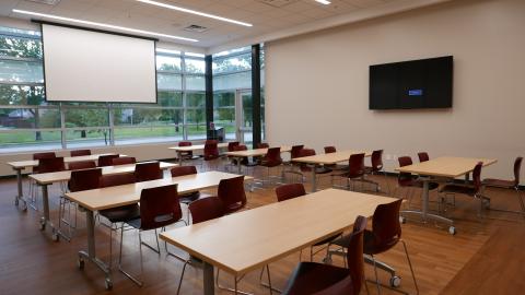 Meeting Room B with 8 rectangular tables, seating for 4 at each table, a white screen and a mounted tv