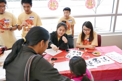 Asian student writing Chinese characters with a paintbrush for mother and child. Other students are eating Chinese food in the background.