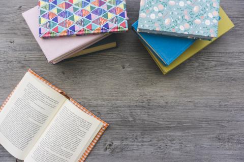 An open book on a wooden background with several more books with colorful covers stacked nearby.