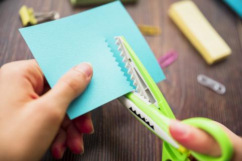 Hands cutting blue paper with green zigzag scissors.