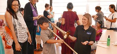 Child playing a trombone with the help of an instructor. People watching in the background.