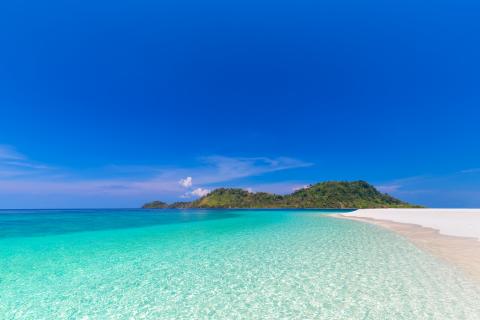 Photo of one of the islands in Hawaii. Blue sky and blue-green water with a sandy beach to the right side. Lush green foliage in the center.