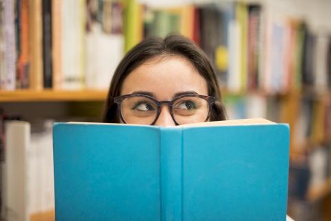 A brunette woman with glassesholding up a blue book that is covering the bottom half of her face.