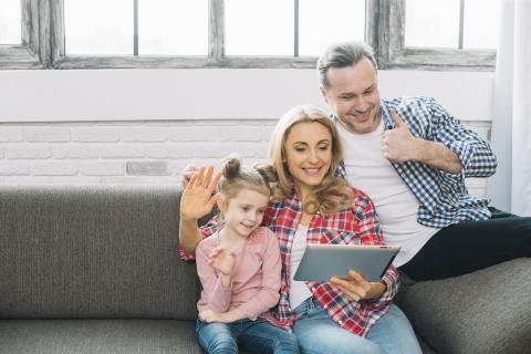 Man, woman, female child sitting on a grey couch looking at a tablet together.