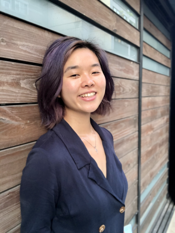 Laura Gao, Asian American author, smiling and leaning against wood paneled wall.