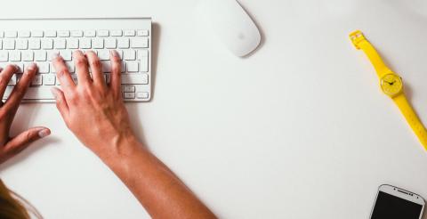 Hands typing on a keyboard with a mouse, yellow watch, and phone to the side.