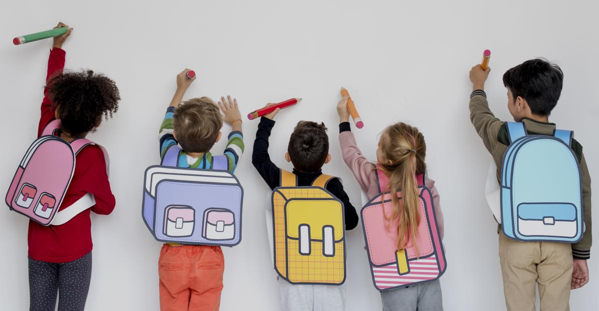 Five children wearing colorful backpacks facing a wall using large pencils to pretend to write on the wall. 