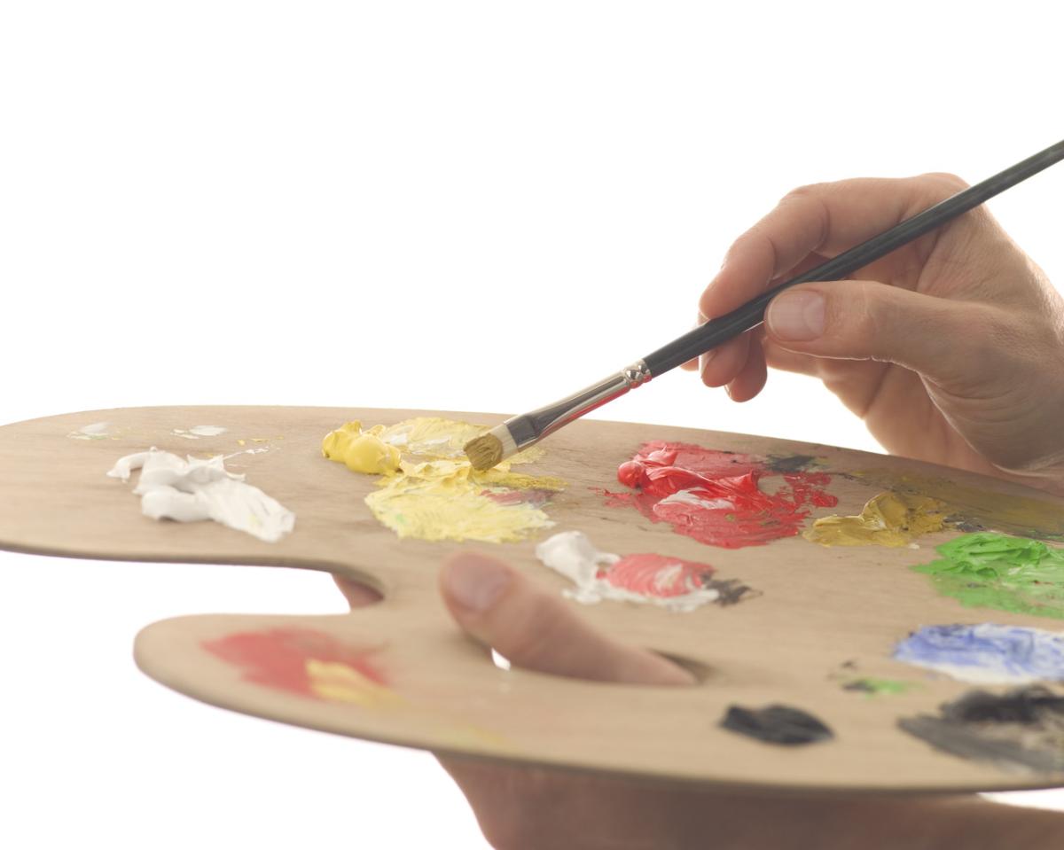 Hand holding a paintbrush and wooden palette with various colors of paint on it.