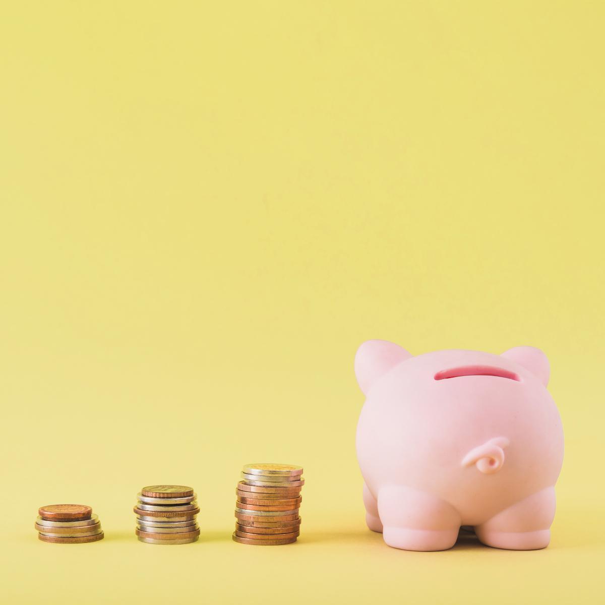 Pink piggy bank facing away from camera with various stacks of coins on a yellow background.