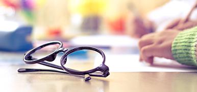 Black framed glasses on a desk with a student writing in the background.