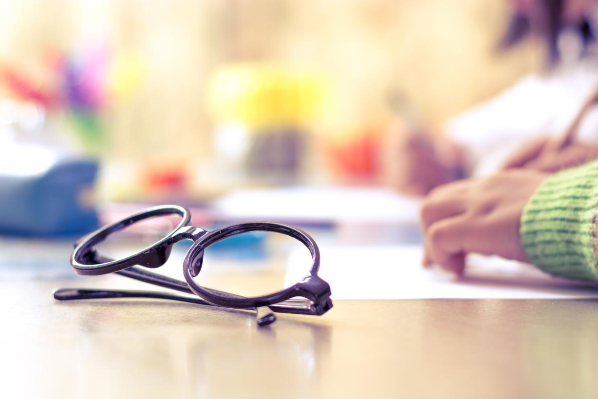 Pair of black framed glasses sitting on a table next to a person's hands writing on paper.