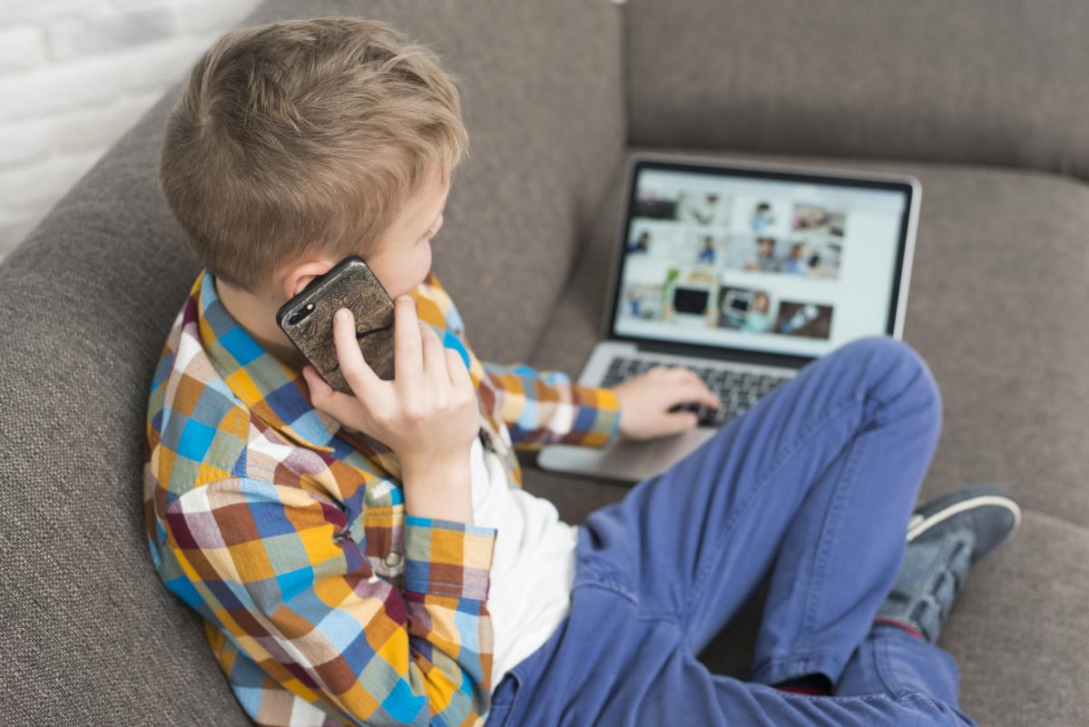 A boy looking at a laptop while using a phone sitting on a couch